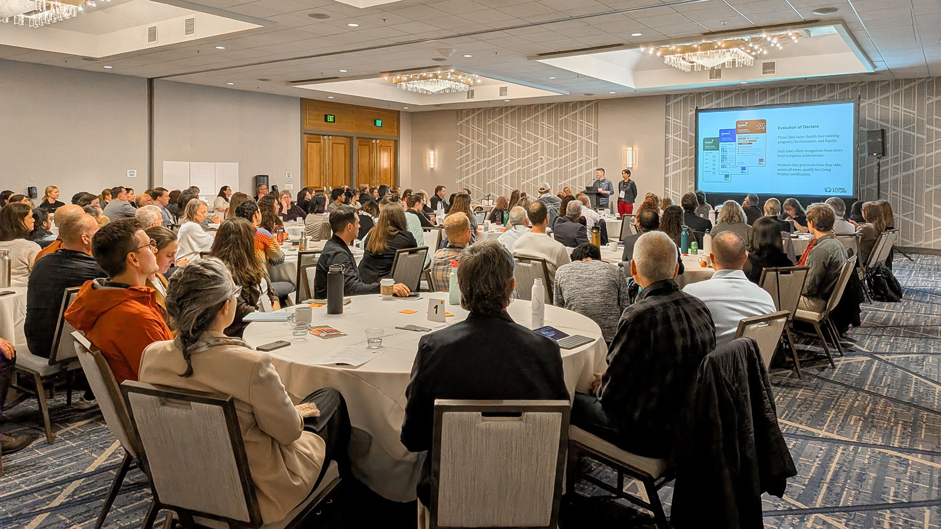 Conference room full of attendees at round tables watching a presentation on a large screen at the front of the room