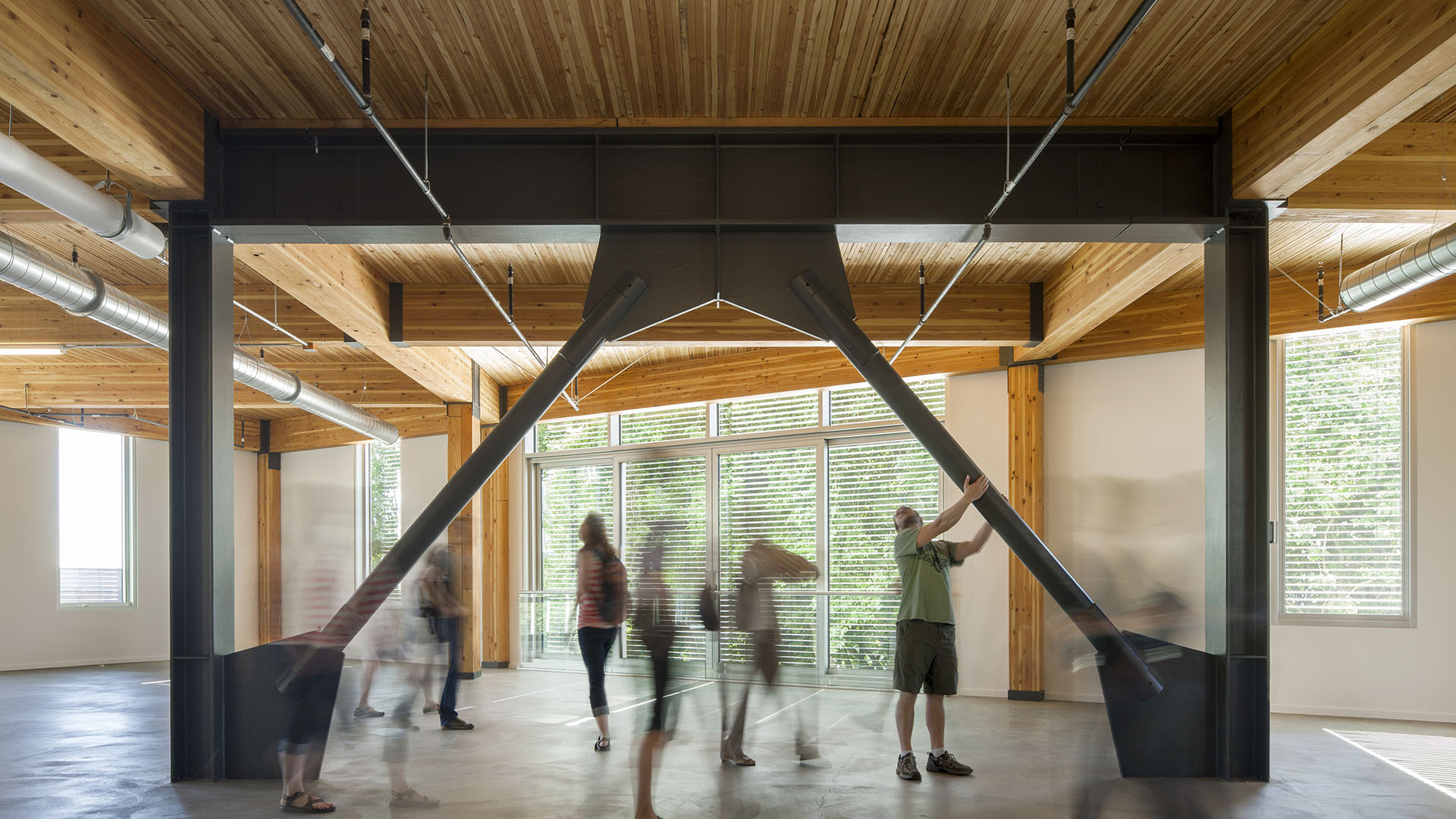 Open interior with exposed wood ceiling and large black x shaped steel braces blurred visitors move through the space