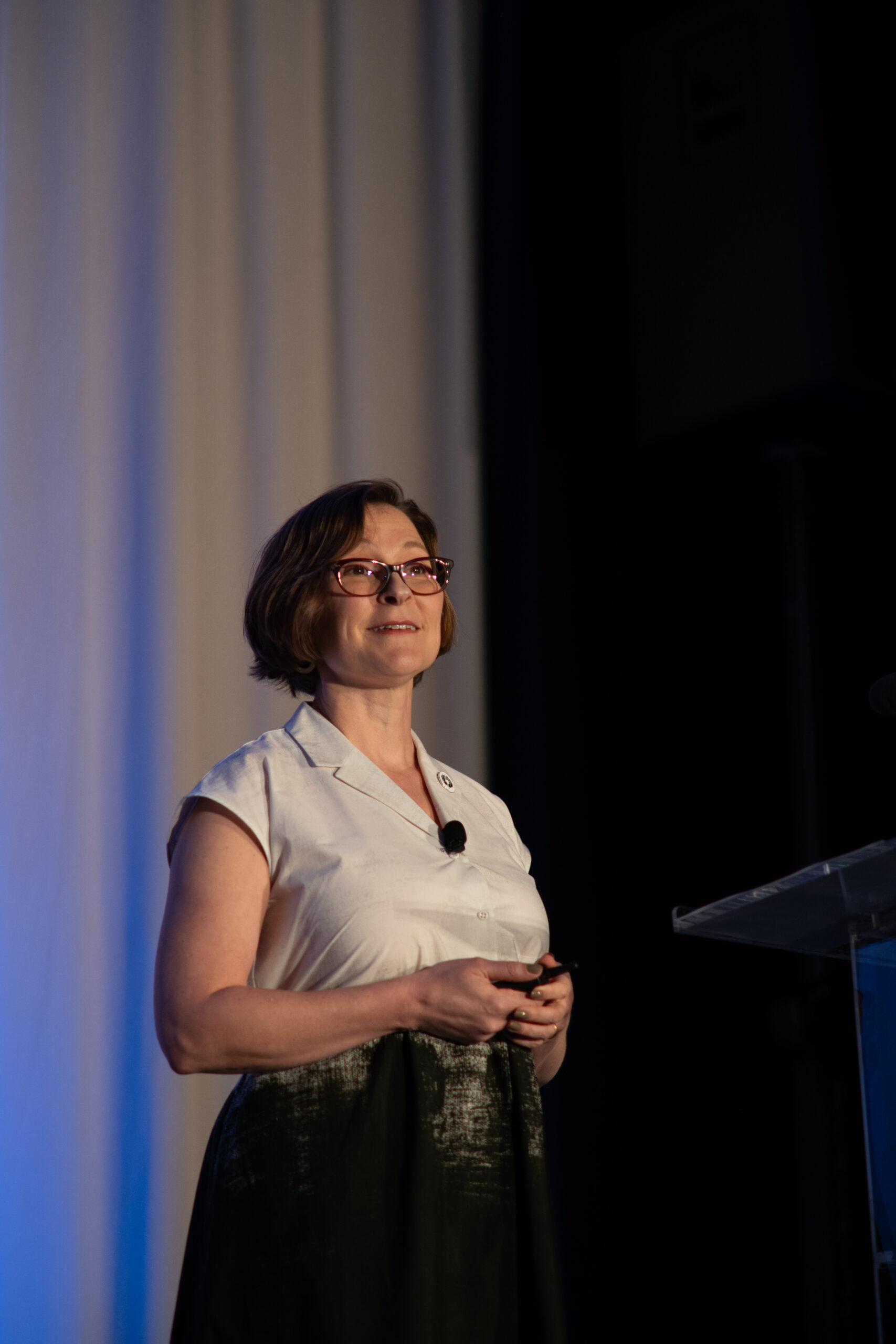 Dsc0676 | trim tab Female presenter with short hair and glasses speaking on stage wearing a light blouse and a microphone headset in front of a blue lit curtain