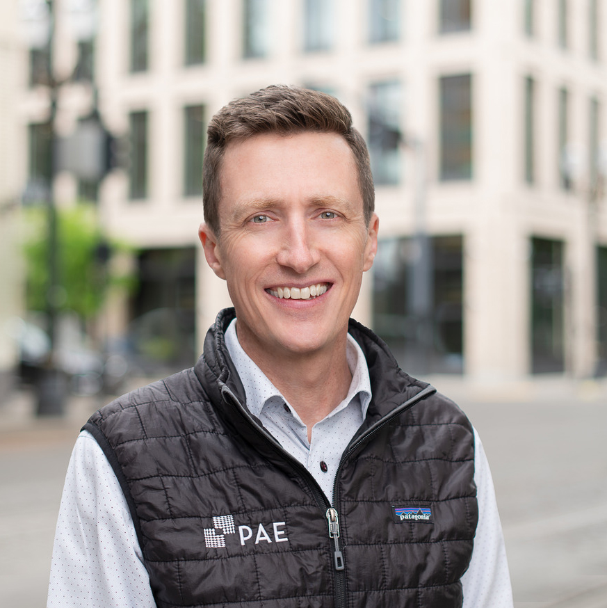 Portrait of a smiling man wearing a black quilted vest with a pae logo over a white dotted shirt standing on a city street There is an urban background with buildings and greenery