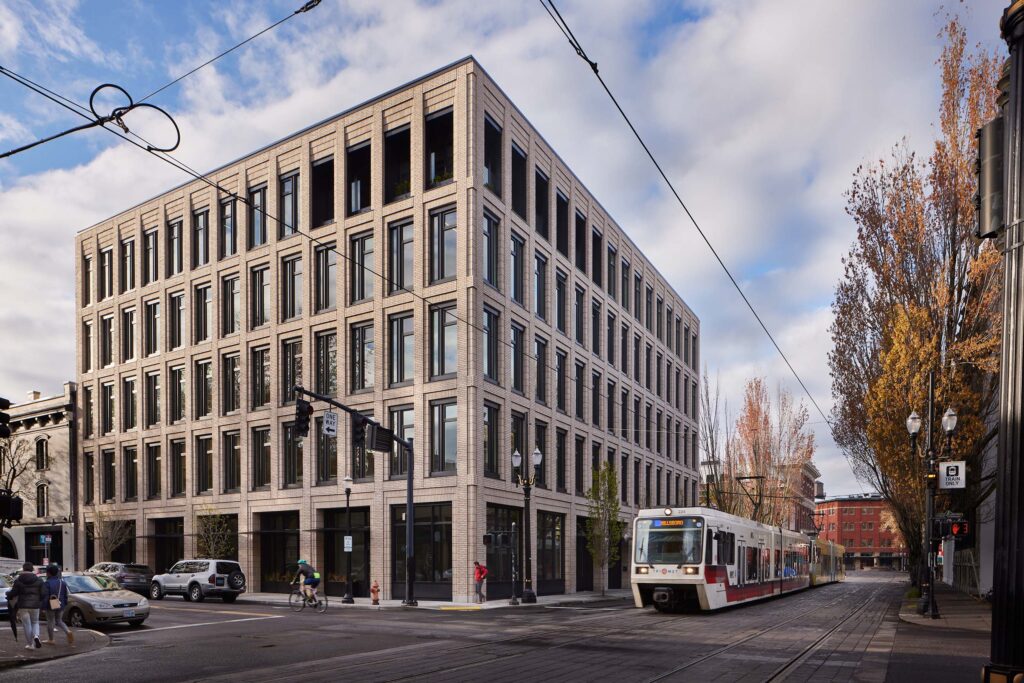 Beige brick office building along a street with a tram on tracks and pedestrians nearby autumn trees in the background