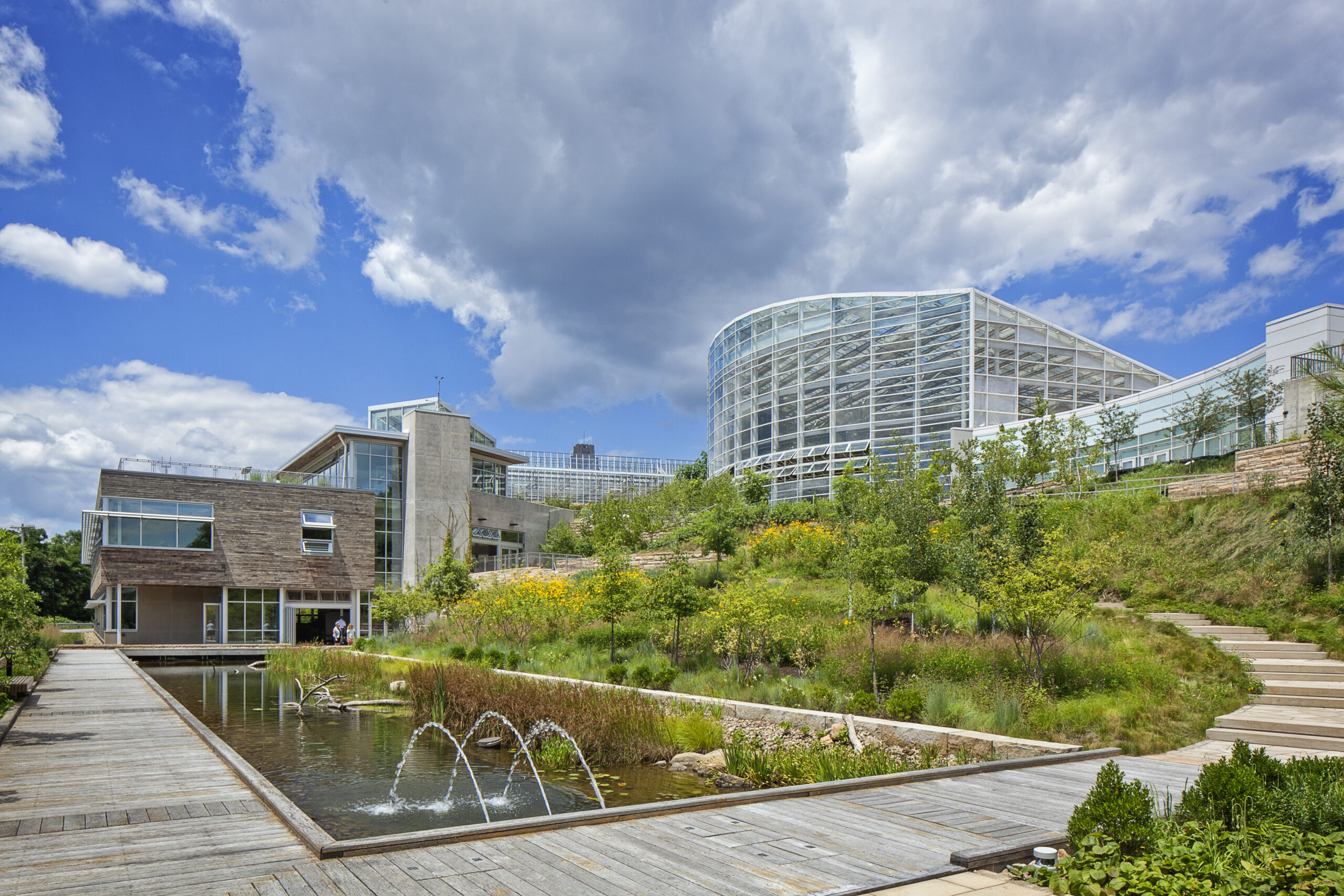 Modern glass domed building atop a landscaped slope with a wooden deck and water feature under a blue sky