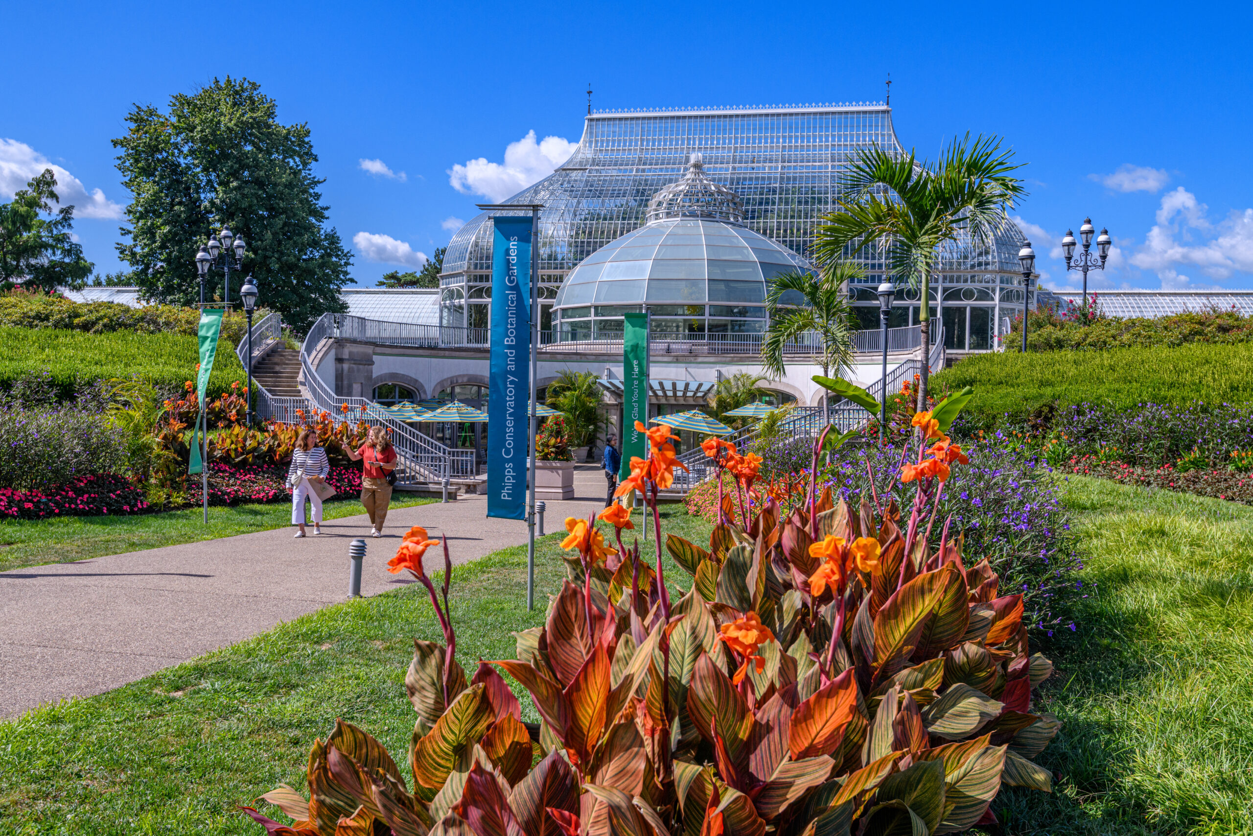 Visitors walk along a paved path beside a glass conservatory with domed sections colorful flowerbeds in the foreground and lush greenery under a bright blue sky