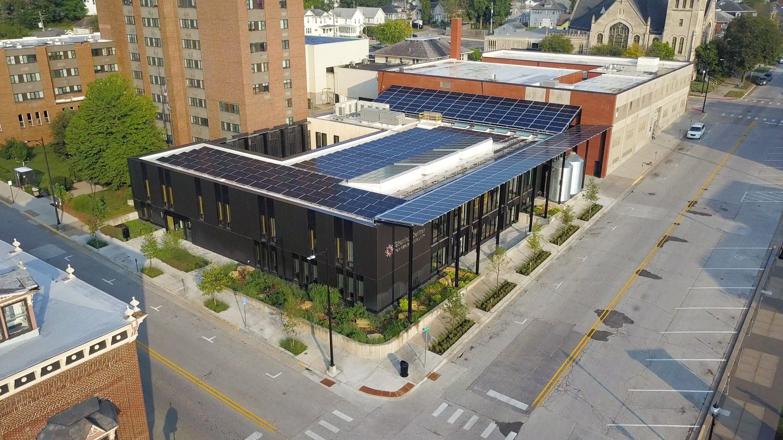 Aerial view of a modern black building with extensive solar panels on its roof amid surrounding brick buildings and streets