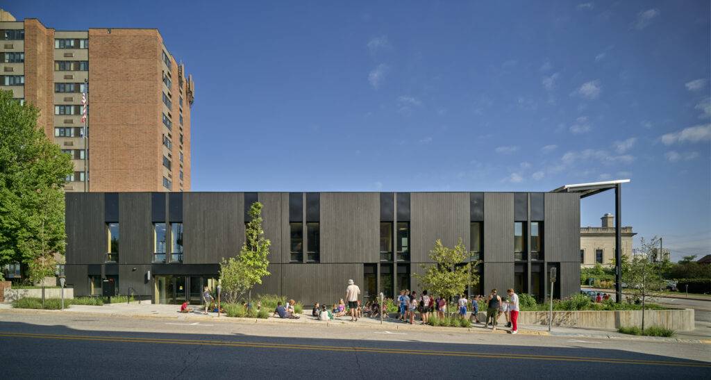 Street view of a black rectangular building on a sunny day A group of people are outside about half are sitting and half are standing near trees and plants