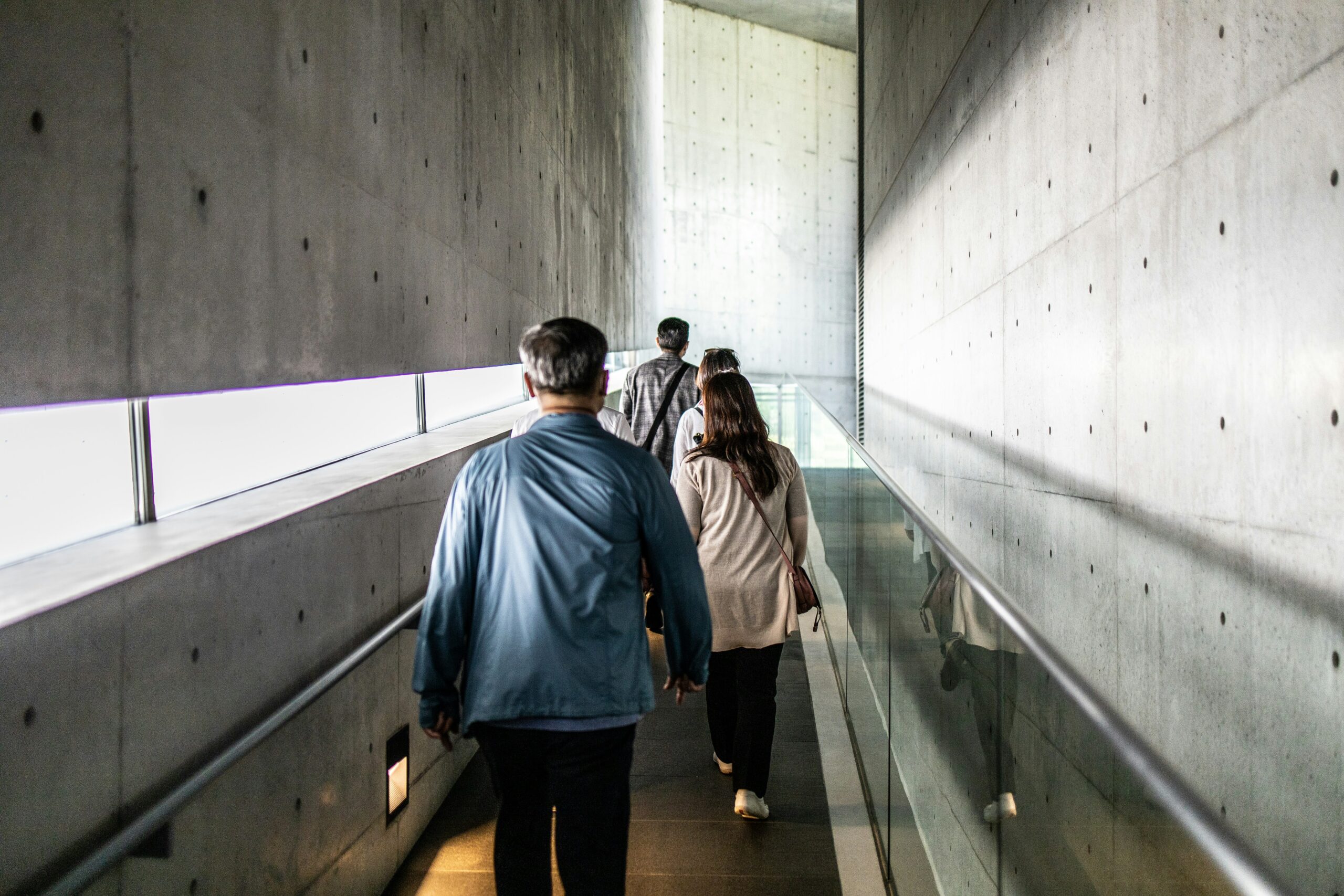 People walking away along a narrow elevated walkway with concrete walls and glass railing seen from behind