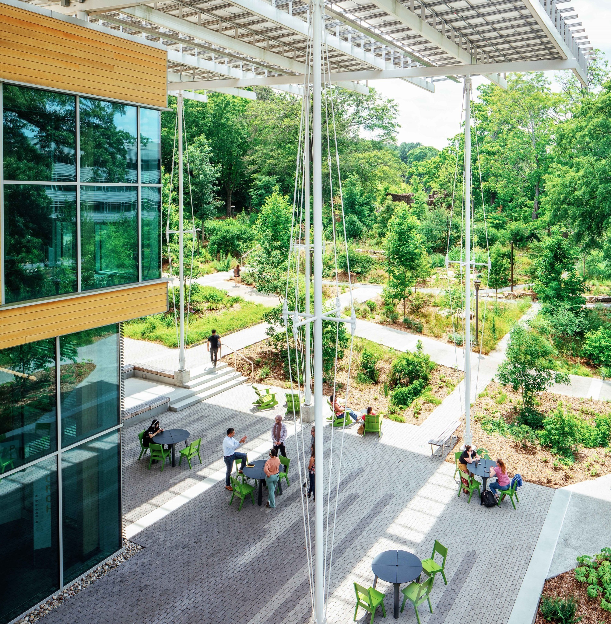 Outdoor patio beside a glass fronted building with lime green chairs around gray tables and overhead metal framework surrounded by trees