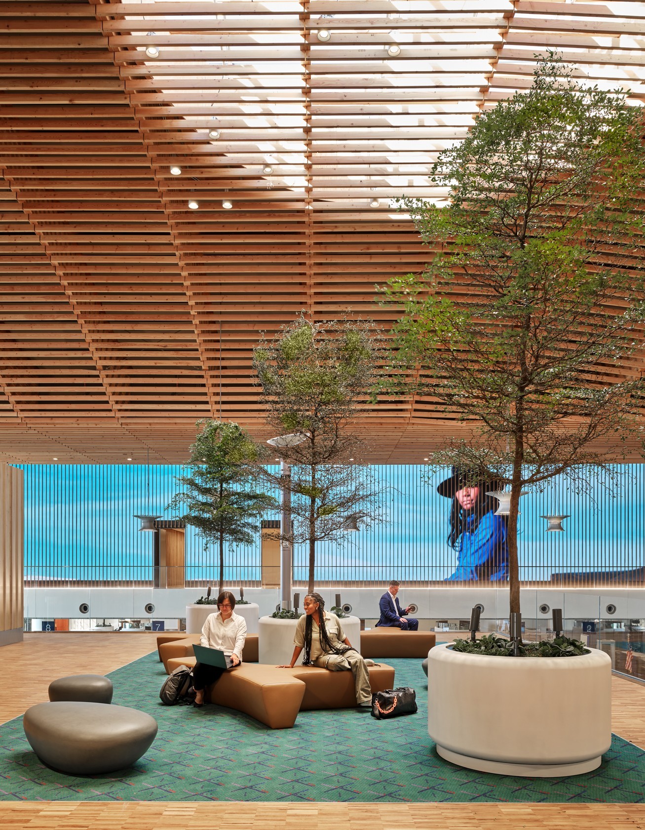 Indoor lounge with a wooden slat ceiling tall potted trees and modular brown seating with people sitting and working nearby